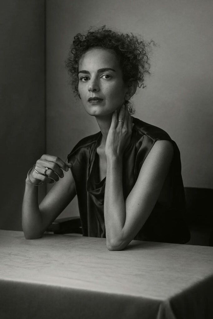 Leïla Slimani, siyah-beyaz portre fotoğrafında ciddi bir ifadeyle objektife bakıyor. Masanın başında oturmuş, elini boynuna götürmüş. Goncourt Ödüllü yazarın güçlü ve zarif portresi.