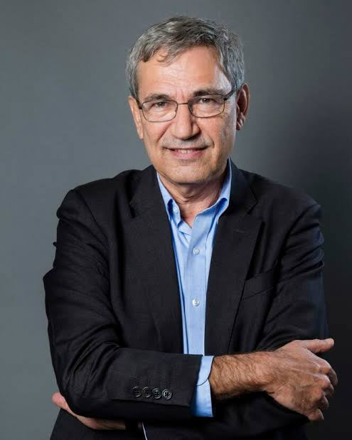 Orhan Pamuk takım elbiseyle, kollarını kavuşturmuş şekilde gülümsüyor.