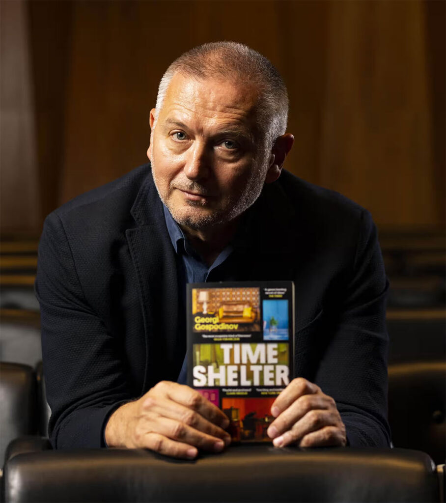 Georgi Gospodinov elinde Zaman Sığınağı (Time Shelter) kitabını tutarken kameraya bakıyor.